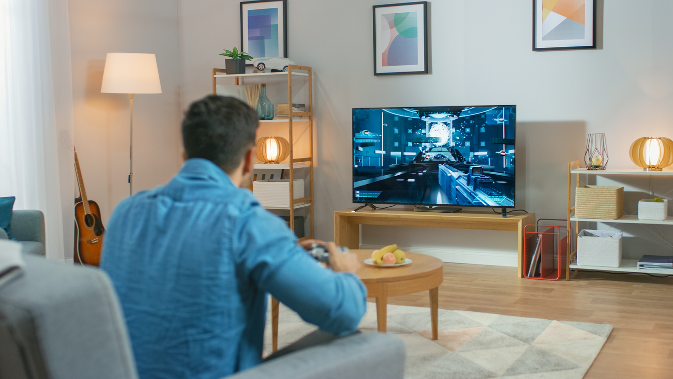 Man Sitting on a Couch Holds Controller Playing in a Console Video Game, 3D Action Shooter Gameplay Shown on TV Screen.